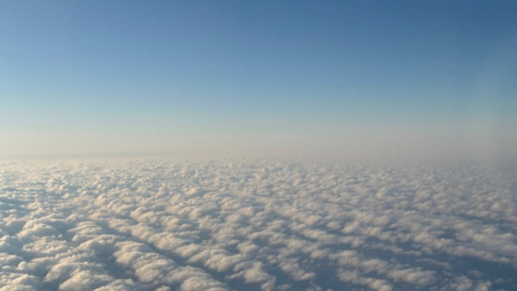Rows of small puffy clouds under a bright blue sky are seen from an aircraft above.