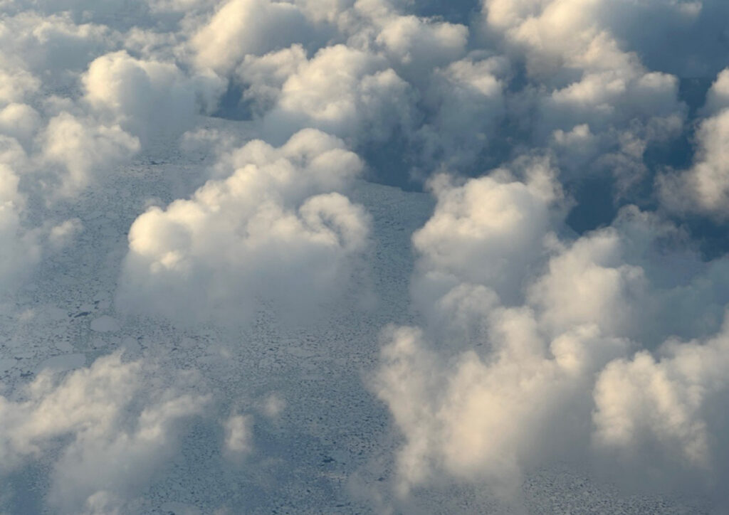 Sea ice on the ocean surface is visible from high above through puffy white clouds.