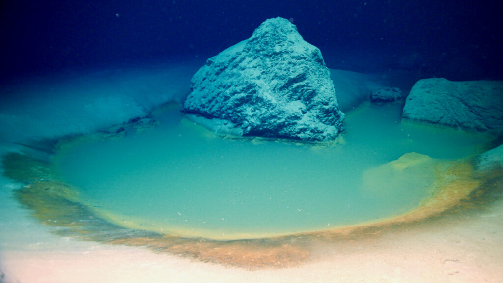 A pool of hazy blue water, surrounded by a ring of darker sand, is seen on the seafloor.
