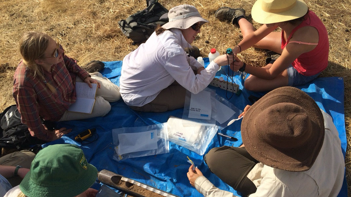 Five people sit on a tarp on a landscape of brown grass. Between two of them is a soil core, and scattered across the tarp are labeled Ziploc bags to hold samples.