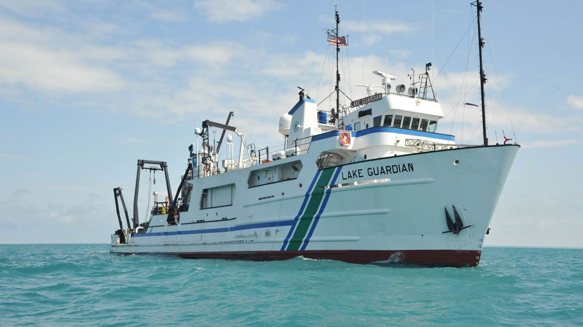 A large ship floats on a body of water on a sunny day. The name of the ship, painted on its bow, is Lake Guardian.