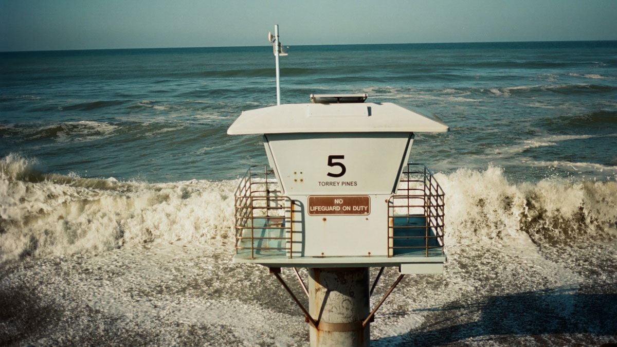 Waves wash up around the base of a lifeguard tower that says “5 Torrey Pines. No lifeguard on duty.”