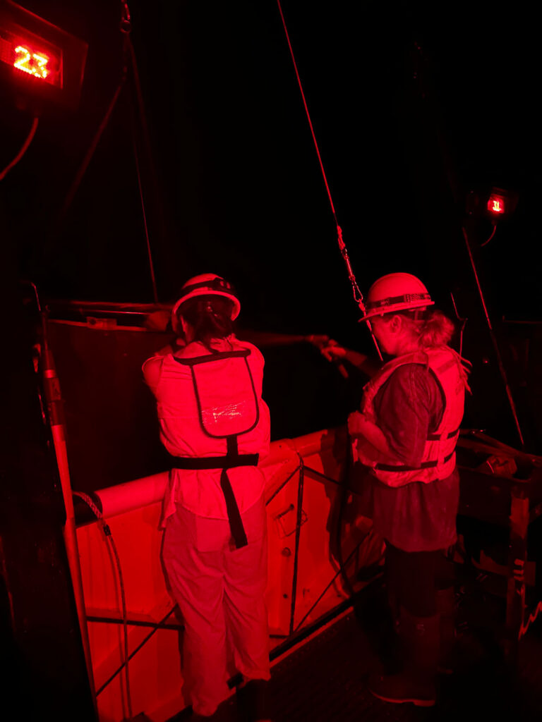 Under red light, two scientists wearing life jackets and hard hats look into a sampling net.