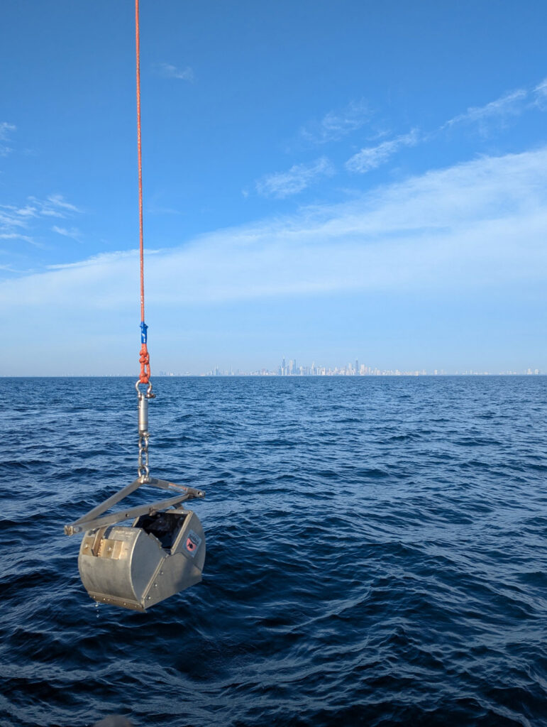Sampling equipment shaped like a metal claw dangles above blue water. The Chicago skyline can be seen in the distant background.