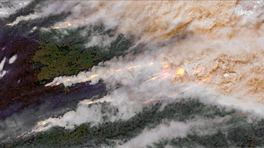 Aerial satellite image showing a large forest fire. The top right of the image is obscured by gray-brown smoke. Several lines of fire extend horizontally across the image.