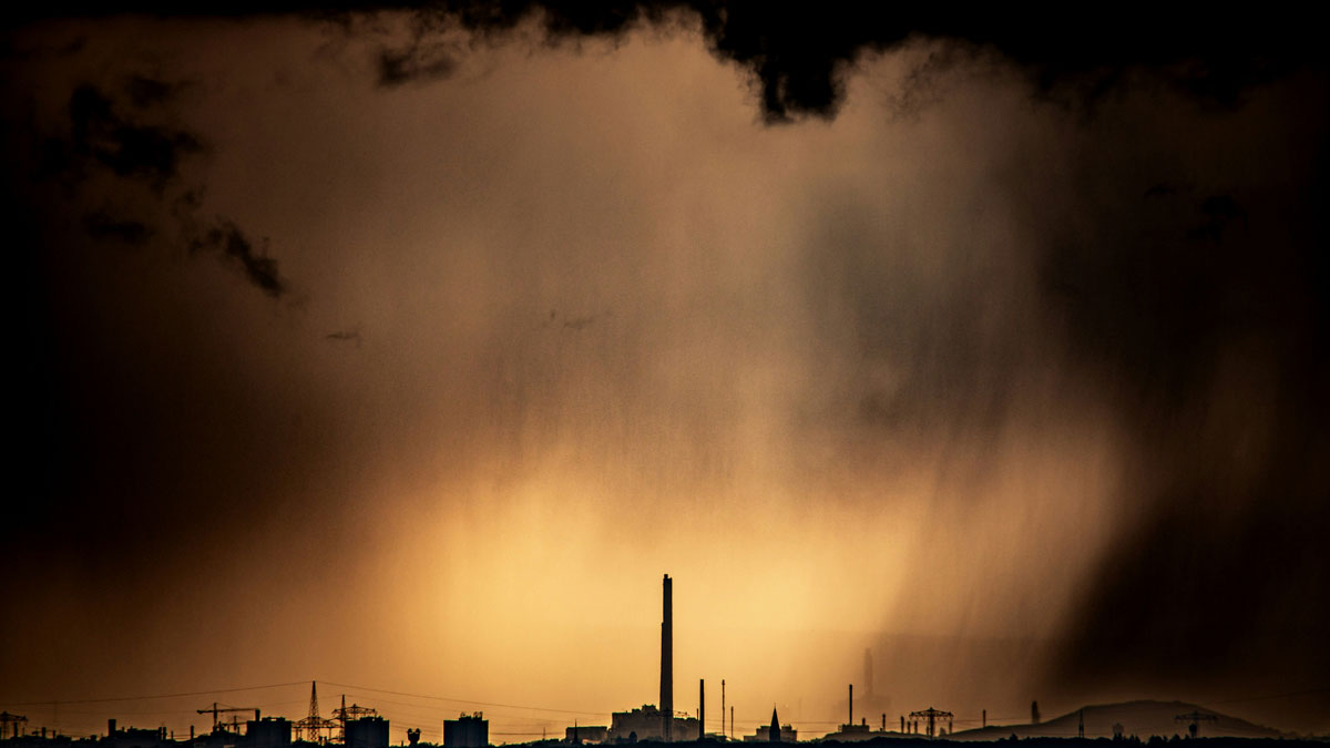 A series of buildings, including several structures that appear to be smokestacks, are silhouetted against a brown sky. Dark clouds are stretched above the buildings.
