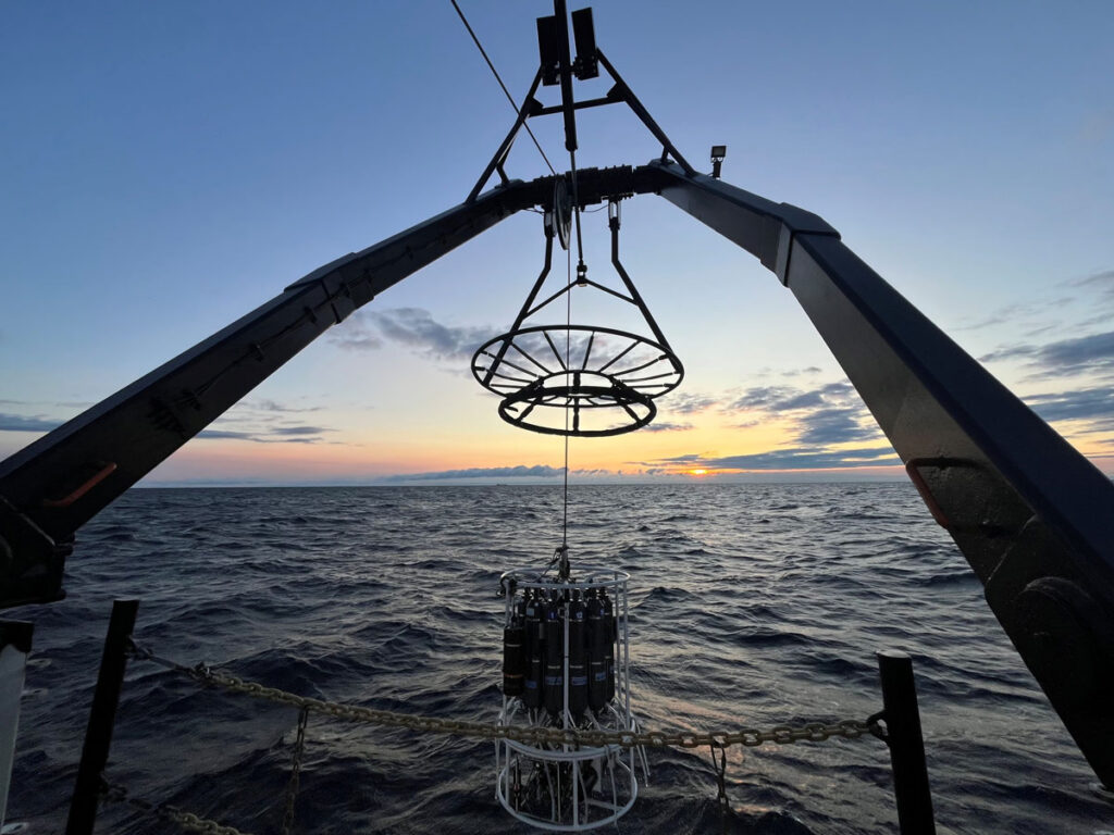 A piece of metal sampling equipment dangles from a metal frame over a lake at sunset.
