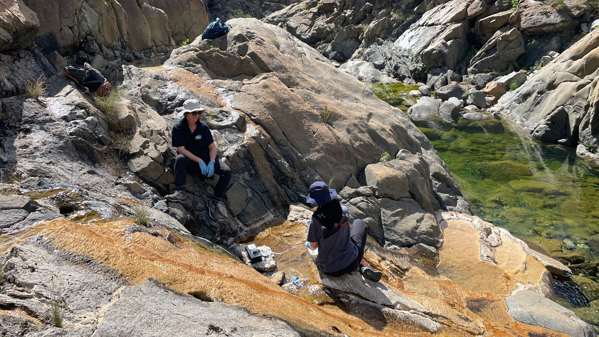 Two people wearing hats and blue lab gloves sit on a rocky slope, part of which is stained orange by spring water, above a green-tinted pool of water.
