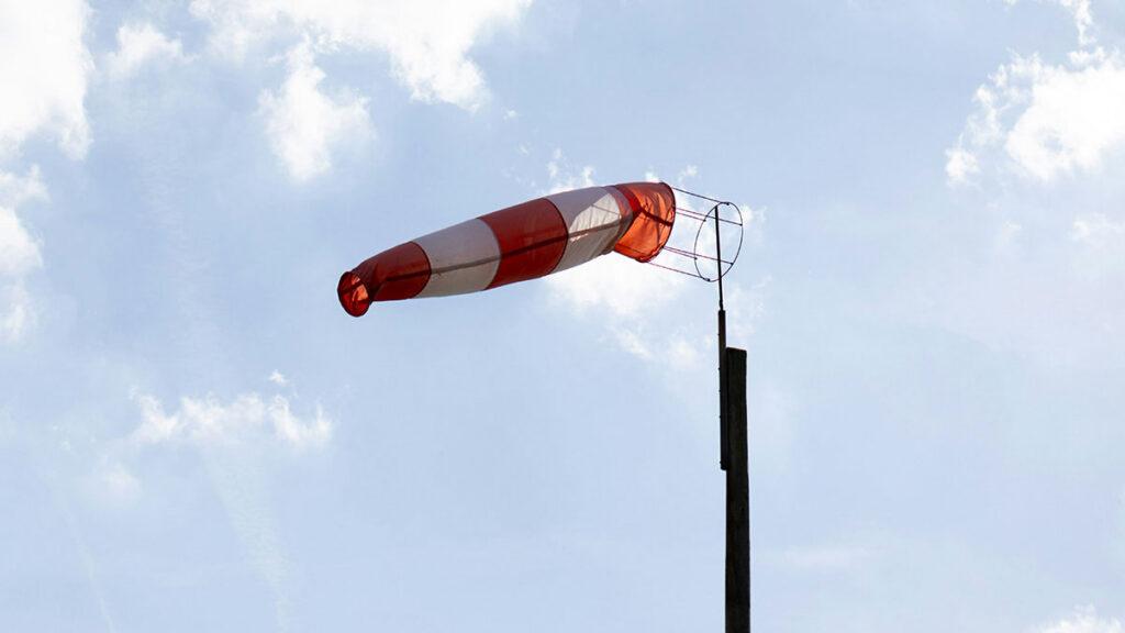A red and white windsock blows in the wind against a blue sky with white clouds.