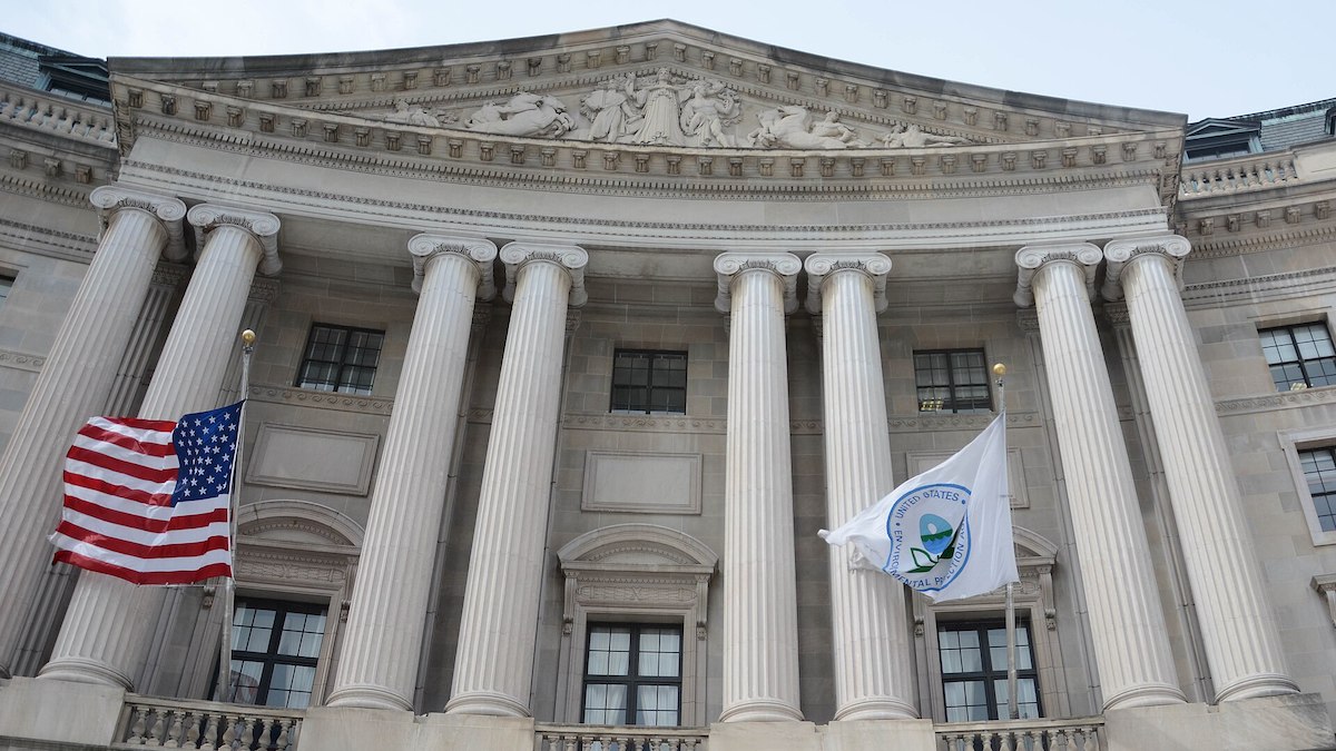 A large, ornate, gray building with columns. Two flags are flying at the building's entrance, the American flag and the EPA flag.