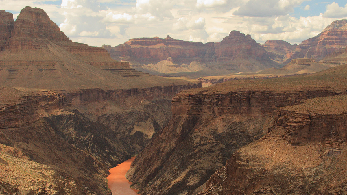 An orange river flows through a canyon. Colorful stratified mesas rise in the distance beneath a cloudy sky.