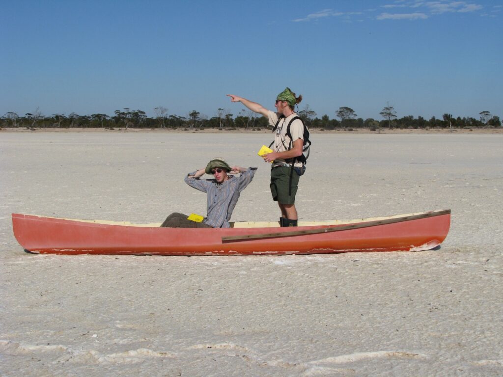Two men are in a red canoe that is sitting on top of a dry lake bed of salt. One has his hands on his head and is looking at the camera. The other stands and points off into the distance.