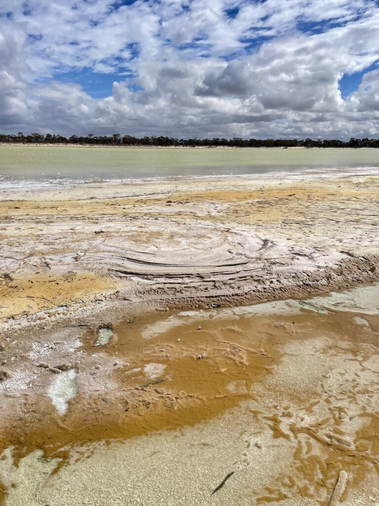 Yellowish shallow water and sand deposits sit beneath a blue sky with clouds stretched across it.