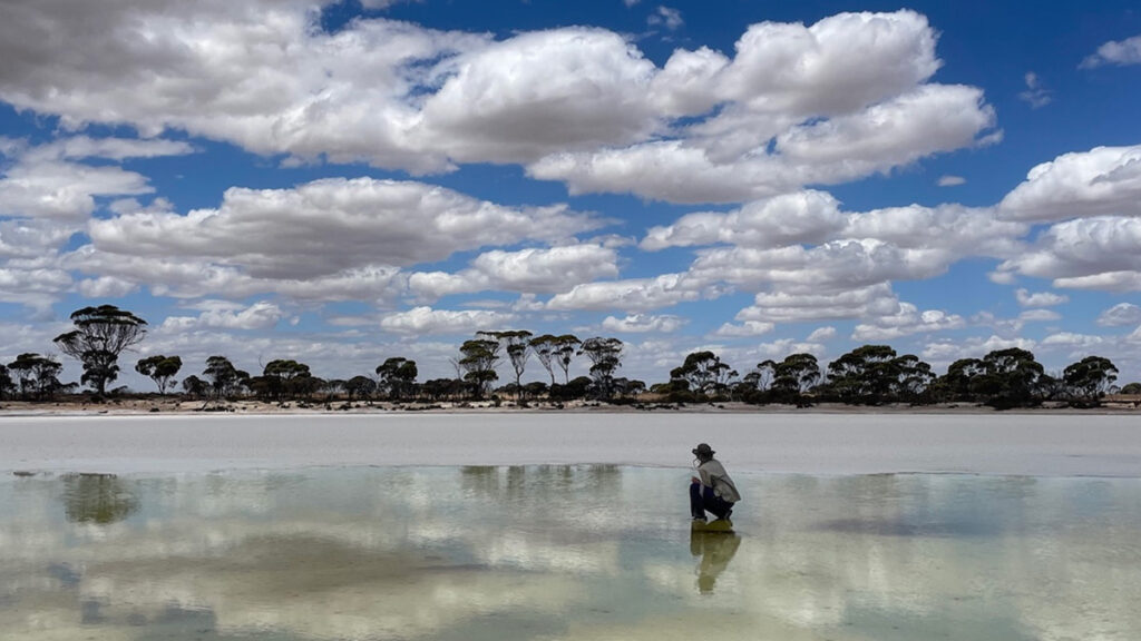 A person takes a knee in the middle of a thin layer of water, which stretches over part of a lake bed. Clouds are reflected on the water’s surface.