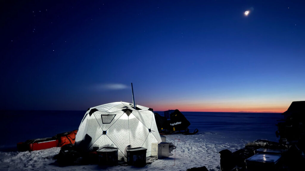 Equipment, including a snowmobile and various crates, surrounds a brightly lit, dome-shaped white tent at night.
