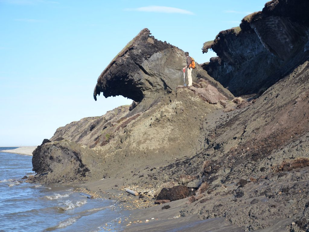 A researcher with a shovel and backpack stands beside a large block of permafrost soil that has separated from the ground behind it and is slumping toward the ocean.