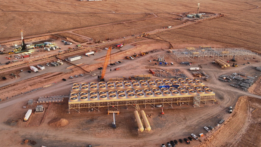 An aerial image shows infrastructure being constructed in the desert, including two towers and a building that appears to be topped with a series of fans. Mountains are visible in the distance.