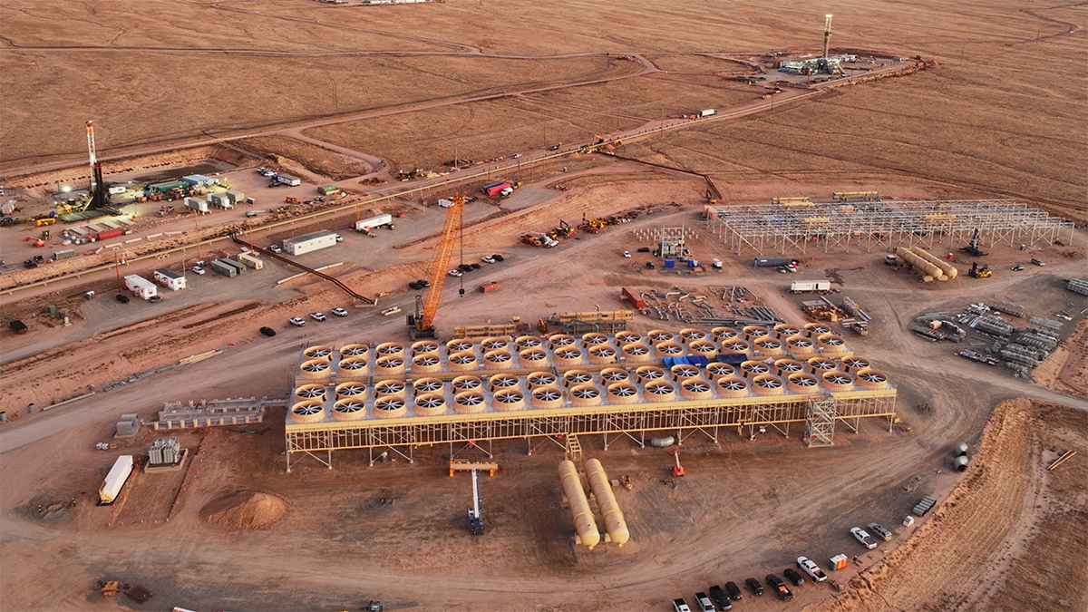 An aerial image shows infrastructure being constructed in the desert, including two towers and a building that appears to be topped with a series of fans. Mountains are visible in the distance.
