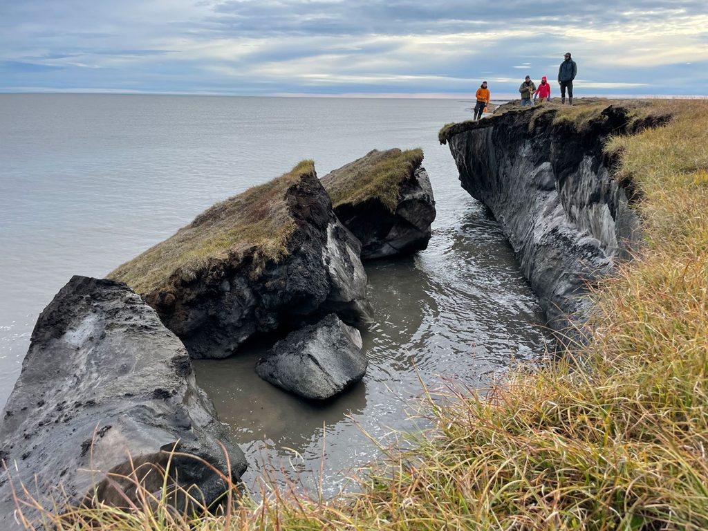 Four people in the distance stand atop ice-rich permafrost bluffs overlooking the ocean. Several massive permafrost blocks that have broken off the shoreline sit nearby in the water.
