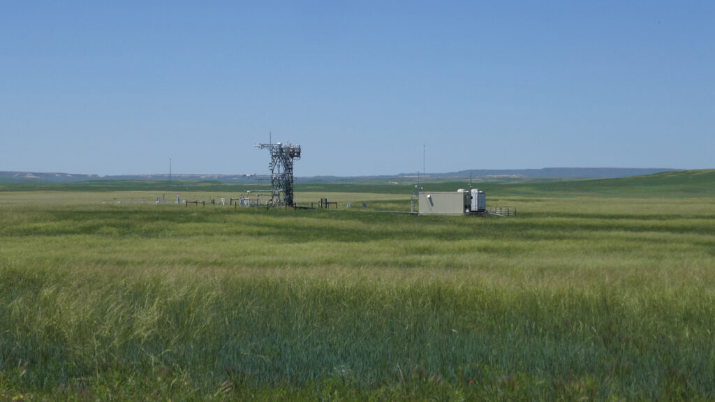 An instrumented metal tower and a metal shed are situated amid a broad grassland expanse under a blue sky.