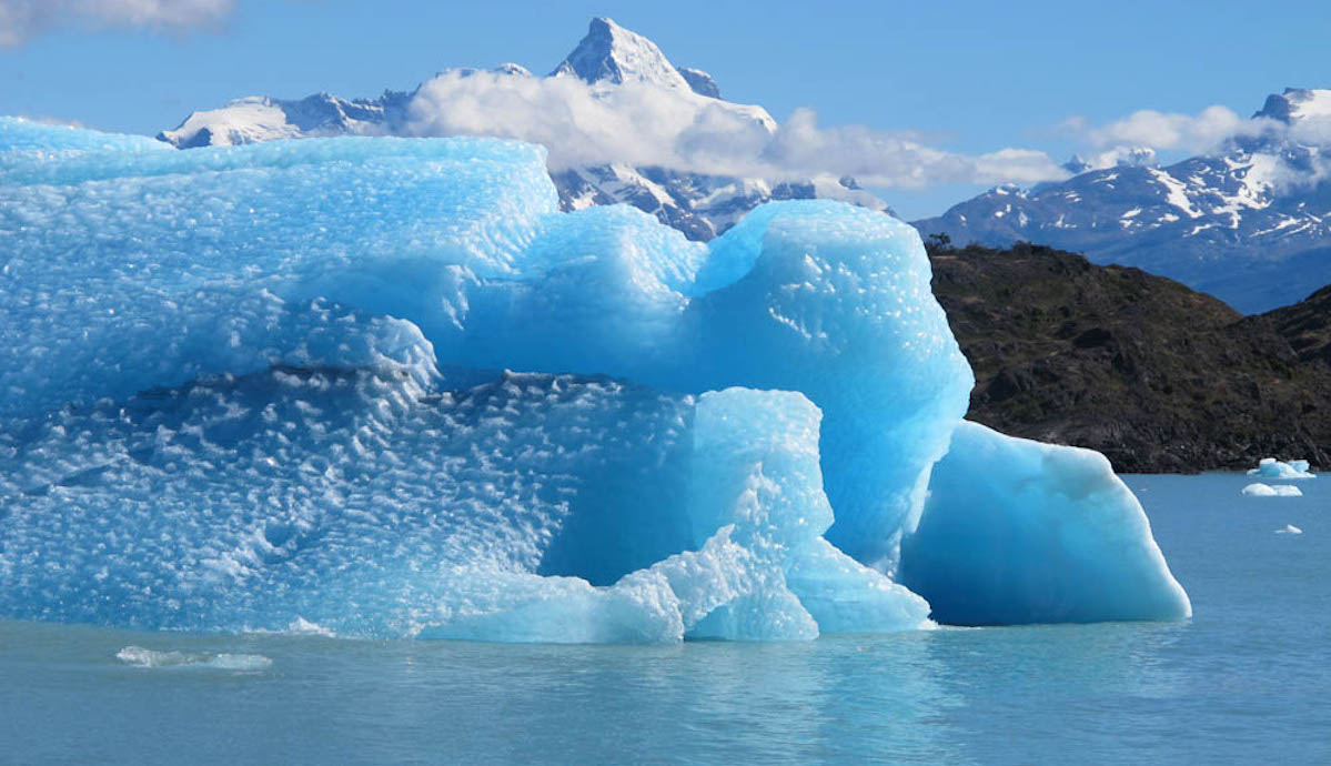 A bright blue chunk of ice, recently calved from a glacier, floats on water with a mountain in the background.