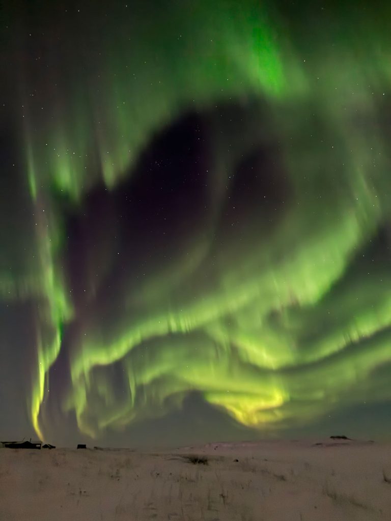 Swirling green and yellow aurorae light up a vast portion of the night sky above a snow-covered expanse.