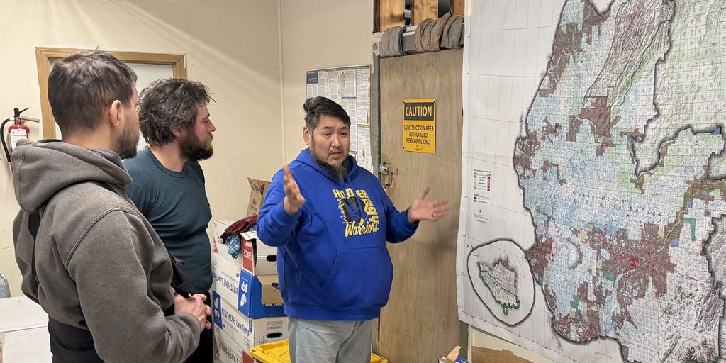 Three people, two of whom are listening to the third speak, stand by a large wall map showing part of western Alaska.