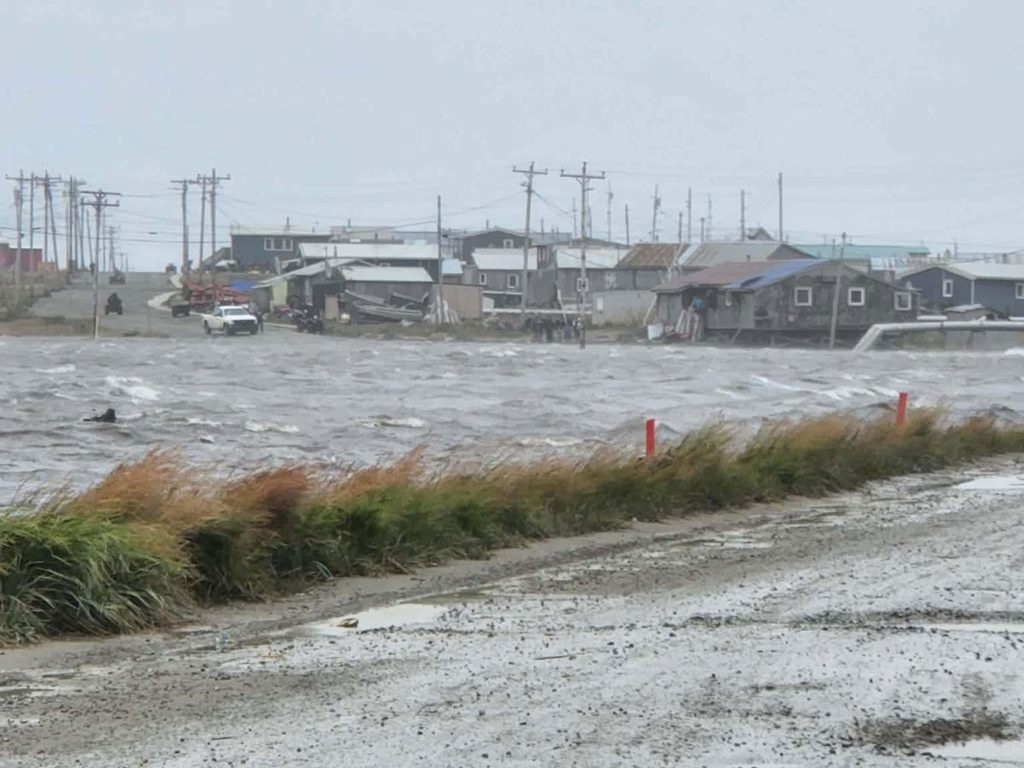 View from a wet roadway across an inlet of choppy water toward a low buildings and homes in the background. Grass alongside the road appears to be being blown by high winds.