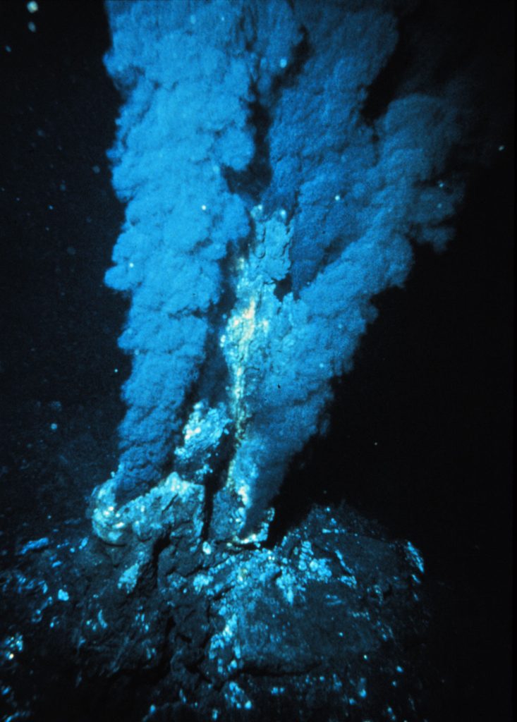 A blue-black image of a vent on the ocean floor releasing plumes of black smoke into the water.