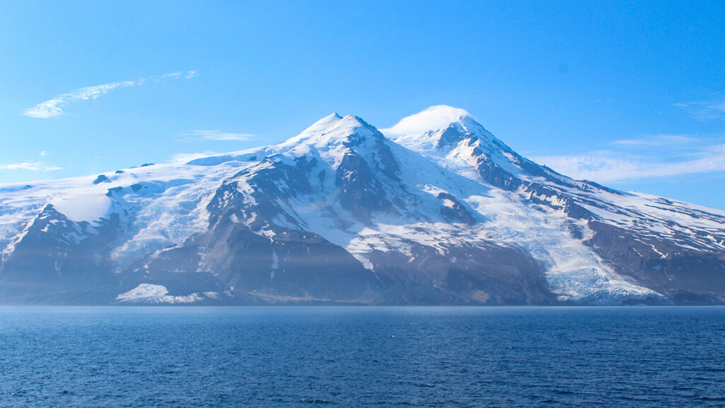 Image of a volcanic island taken from the water. The volcano’s coastal slope has two glaciers. The glacier at right reaches the water’s edge, whereas the glacier at left is covered by rocks near the coast.