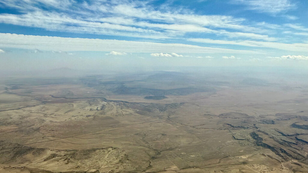 Haze hovers in the distance over an undeveloped, arid-looking landscape as seen from an aircraft. Above, long, thin white clouds stretch across a bright blue sky.