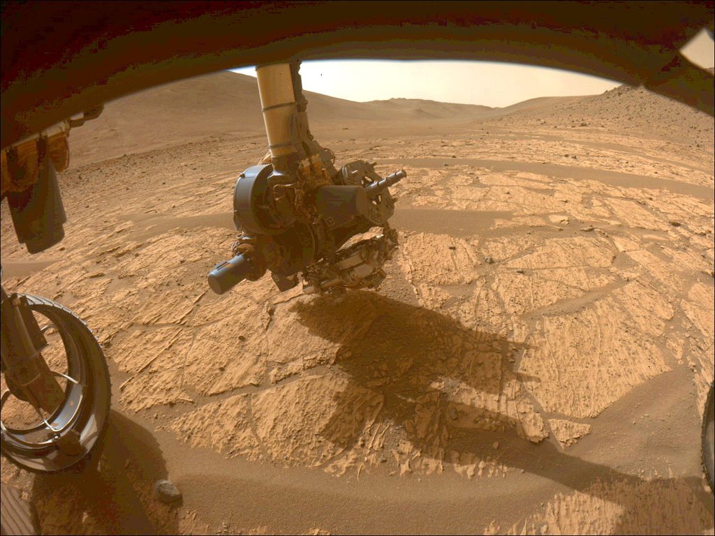 A view from beneath the Perseverance rover showing instruments on the rover’s robotic arm being held very close to the orangish Martian ground surface.