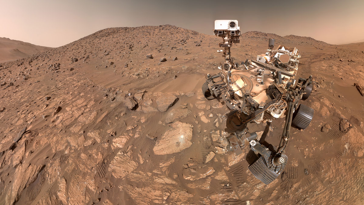 Selfie view of the Perseverance robotic rover sitting on the rocky, dusty surface of Mars, with some low hills and an orange-brown sky behind it.