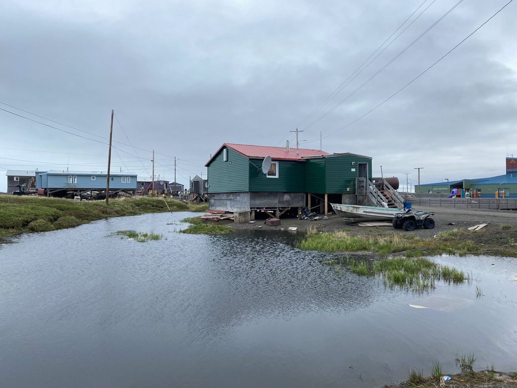 A small house with green siding and a red roof that is elevated on wooden stilts above the ground surface is partly surrounded by standing water filling a depression. A boat hull and a four-wheel all-terrain vehicle sit on the ground in front of the house.