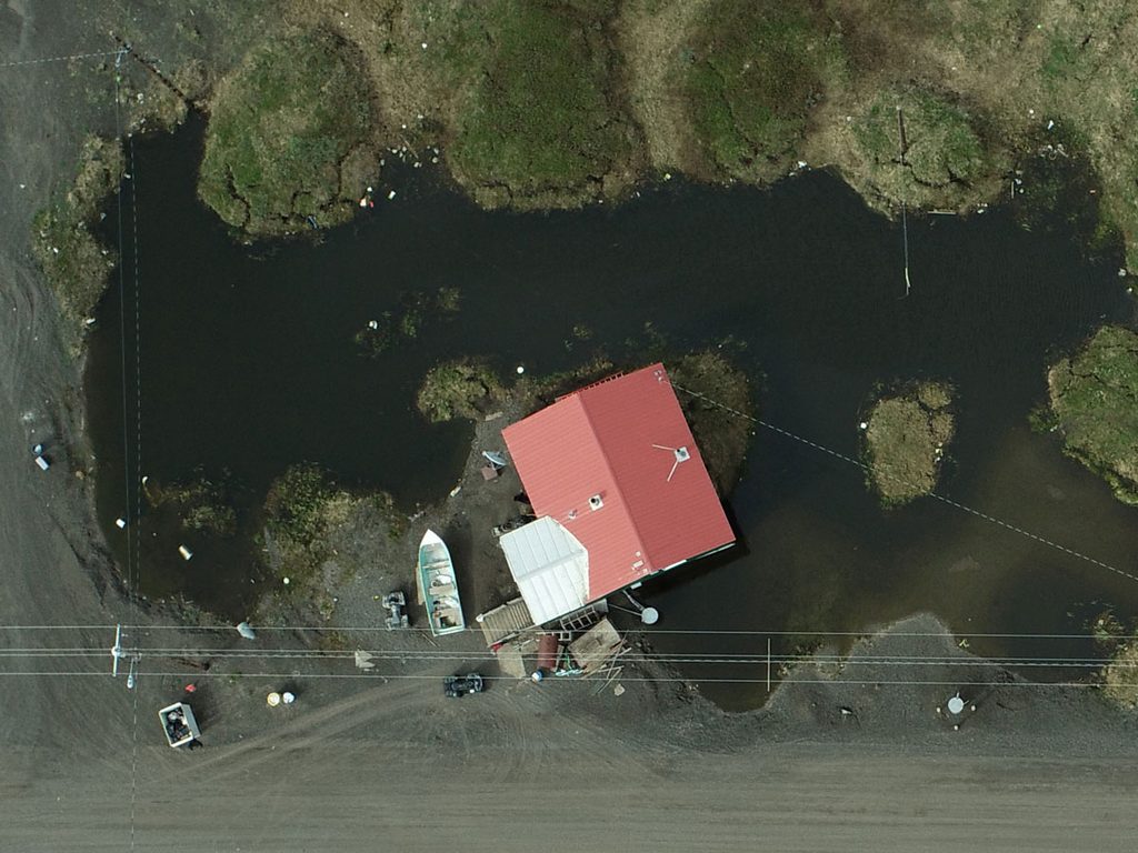 Overhead view of a small house with a red roof that is almost entirely surrounded by standing water.