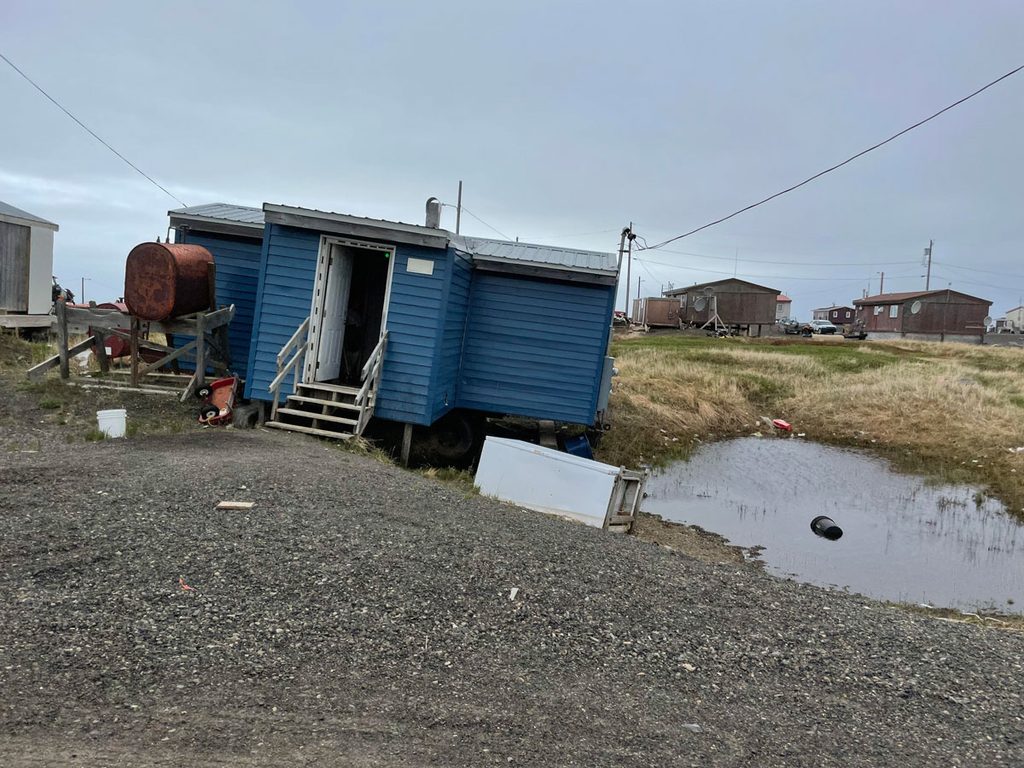 A small house covered with blue siding tilts significantly to one side, toward a water-filled depression in the ground beside it.