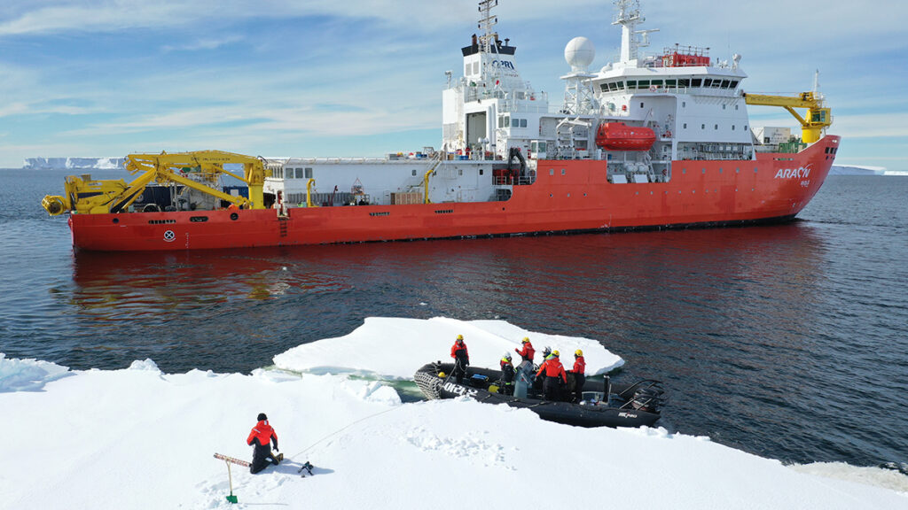 A researcher kneels on a large piece of sea ice to collect measurements as six other people stand in an inflatable powerboat nearby at the edge of the sea ice. A large, orange-hulled icebreaking vessel sits in the open water beyond the researchers in the boat.