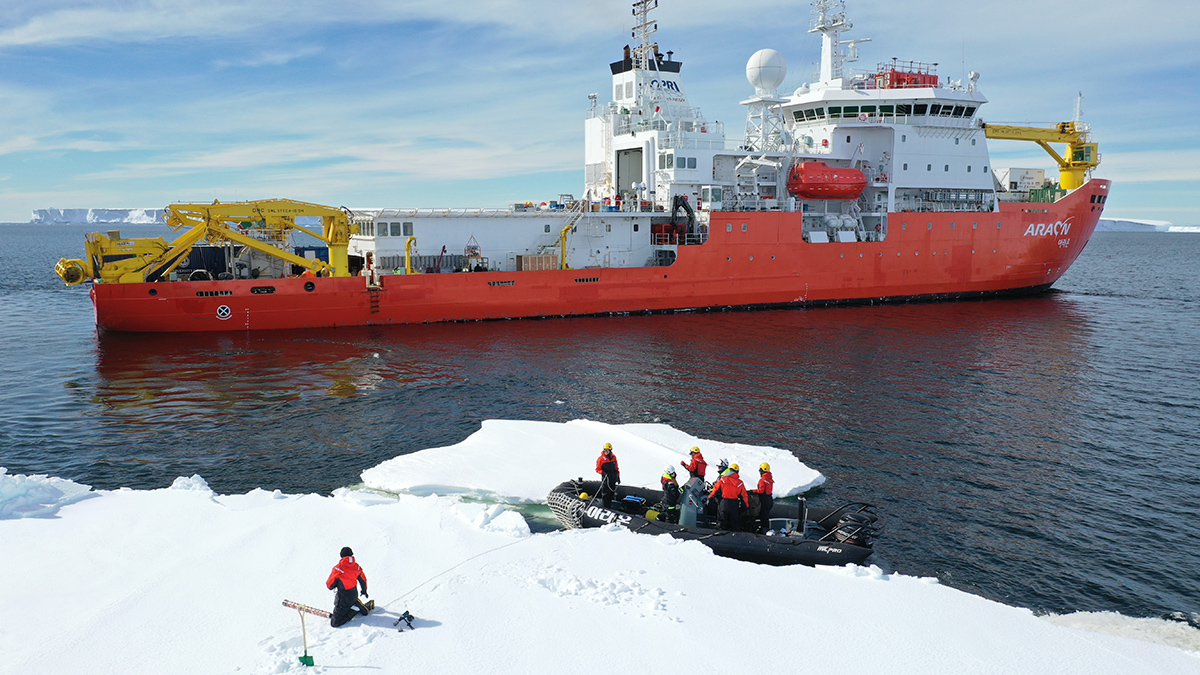 A researcher kneels on a large piece of sea ice to collect measurements as six other people stand in an inflatable powerboat nearby at the edge of the sea ice. A large, orange-hulled icebreaking vessel sits in the open water beyond the researchers in the boat.