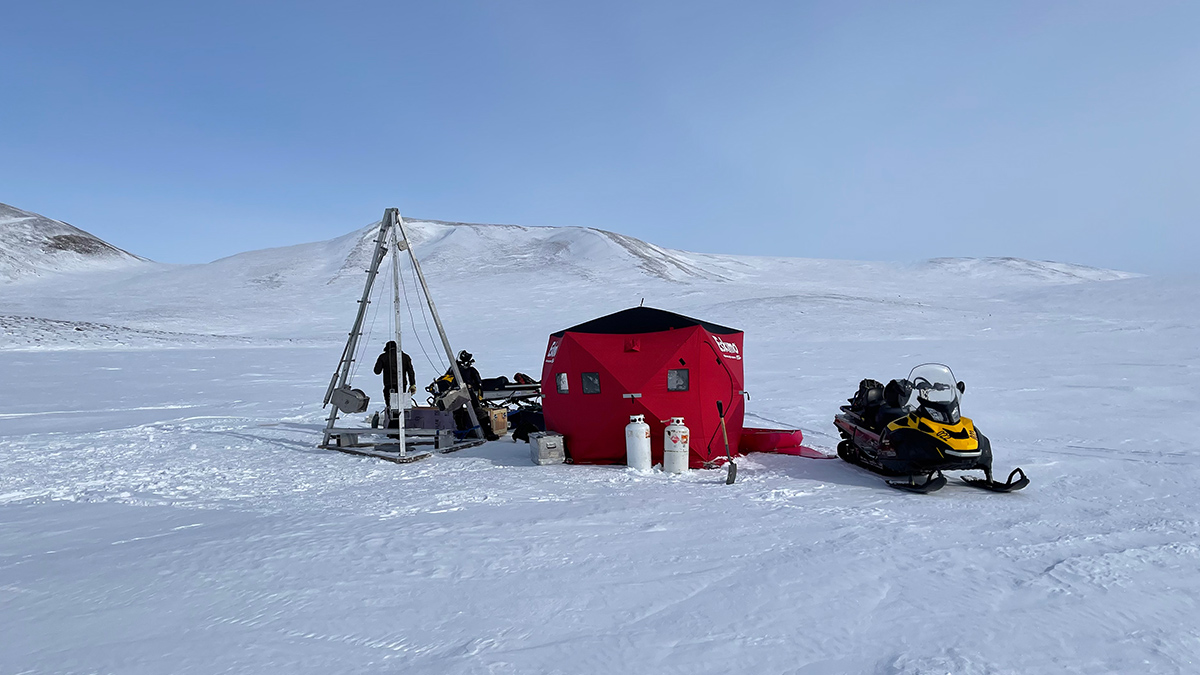 On a snowy, Arctic-looking landscape are a red structure big enough for a few people, a snowmobile, and a large metal tripod structure about twice the height of a human.