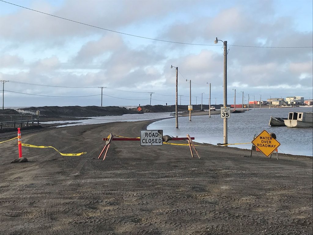 Signage reading “Road Closed” and “Water Over Roadway” blocks a gravel road lined with light poles. A short distance beyond the signs, water covers part of the road, which heads toward a cluster of buildings in the distance.