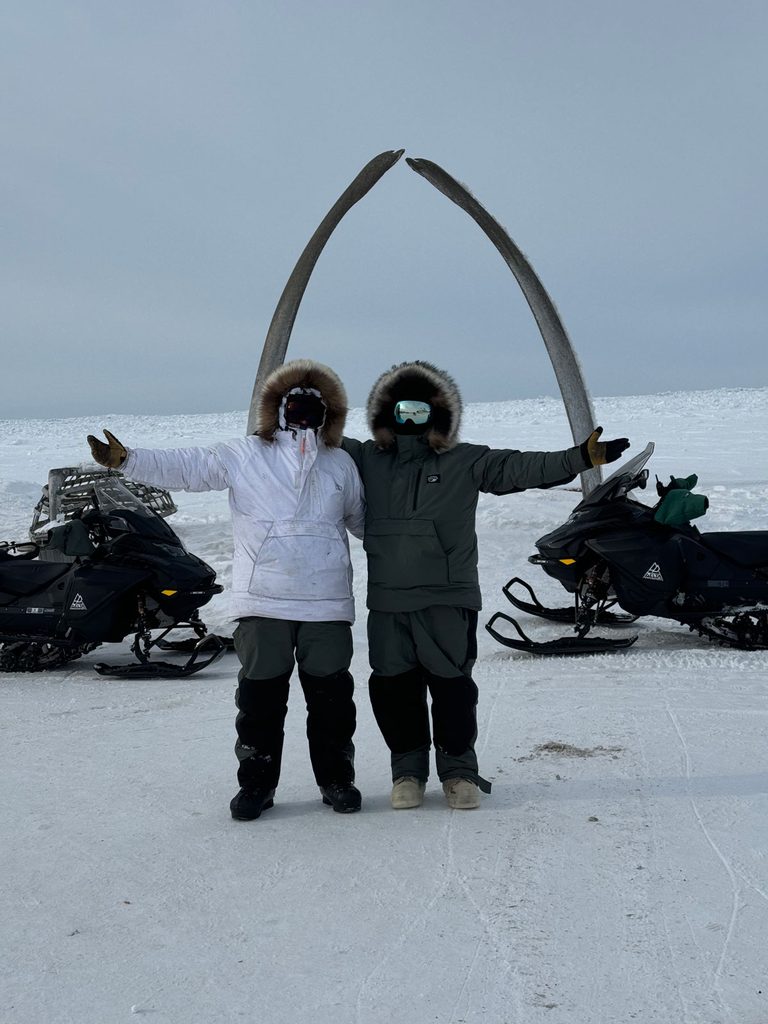 Two people wearing heavy winter clothing and goggles pose for a photo in the snow in front of two snowmobiles and an archway made from two large whalebones.